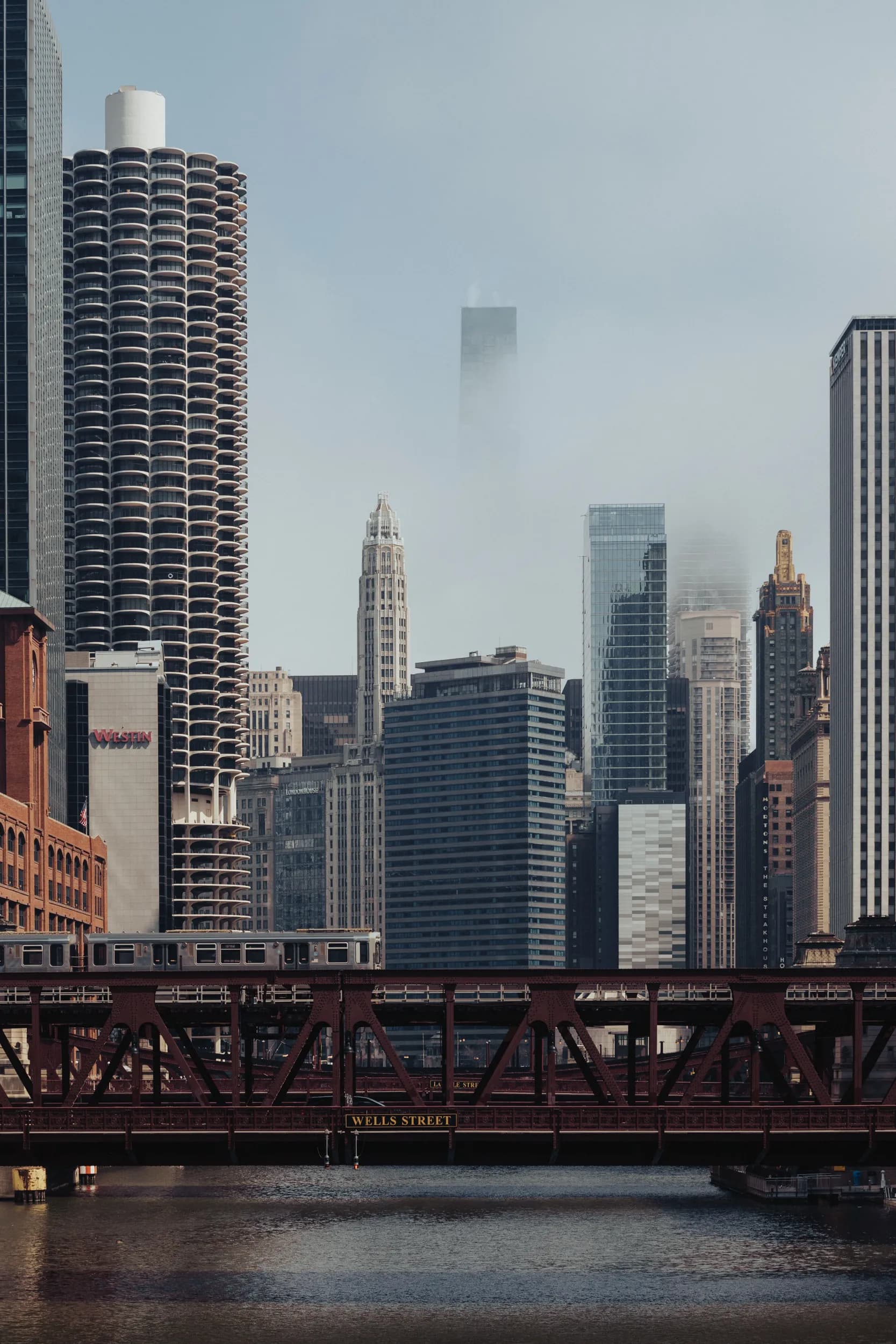 An elevated train crosses a bridge over the Chicago River, with the dense city skyline and Marina Towers rising into the fog.