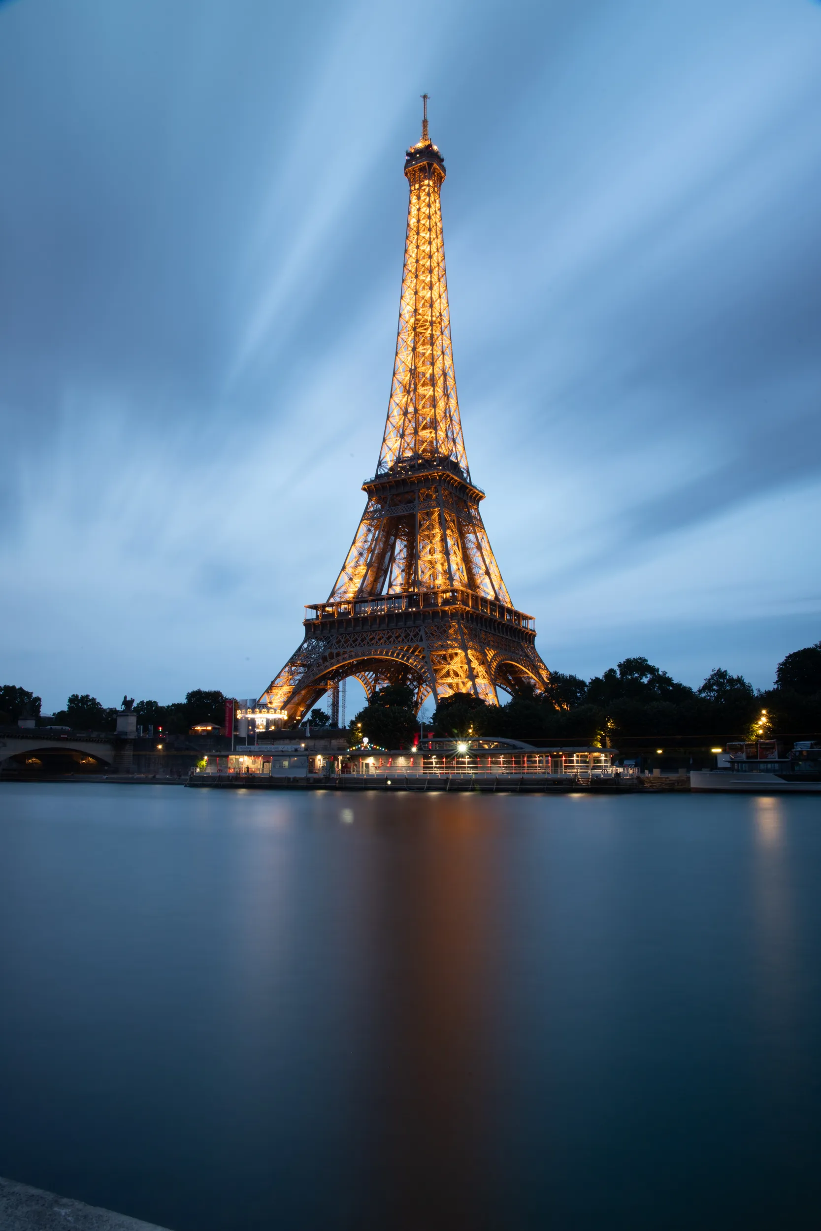 The illuminated Eiffel Tower at dusk, its golden lights reflecting on the smooth surface of the River Seine.